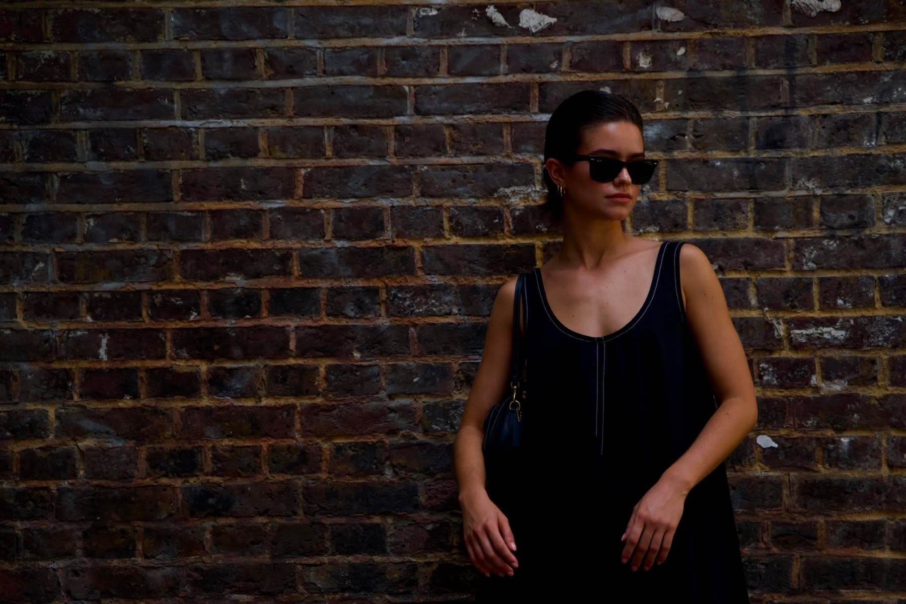 A portrait in a black slip dress and sunglasses against a London brick wall